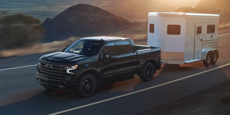Black pickup truck towing a silver trailer on a highway with mountains in the background at sunset.