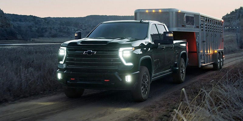 Dark gray Chevy Silverado truck with bright headlights, towing a silver livestock trailer on a dirt road at dusk.