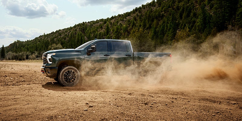 Dark green pickup truck kicking up a large cloud of dust on a dirt trail, with trees and hills in the background.