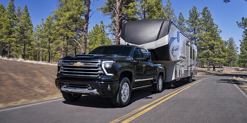Black pickup truck towing a large fifth-wheel RV down a paved road lined with pine trees.