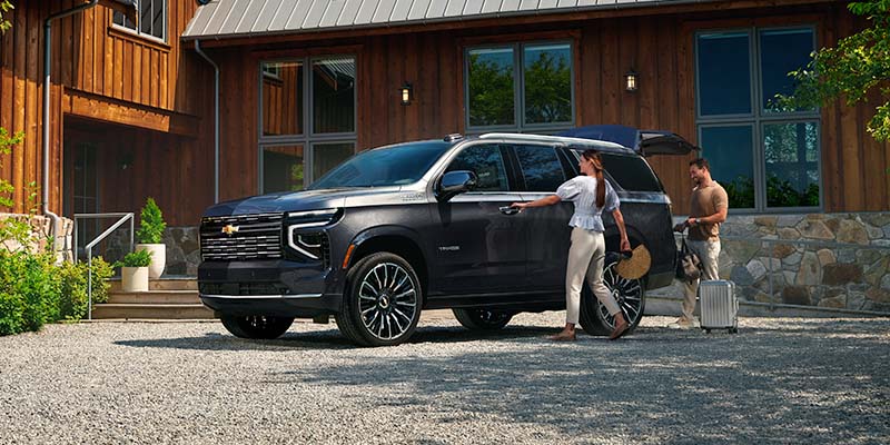Dark grey Tahoe SUV parked on a gravel driveway in front of a modern wooden house, with people loading luggage.