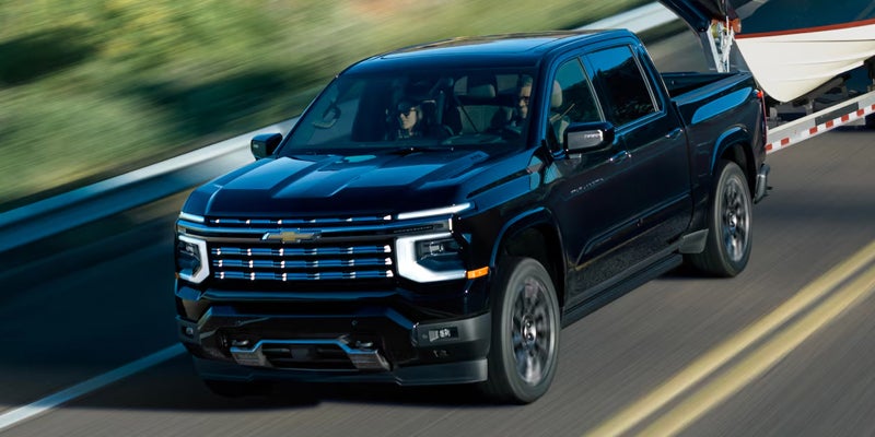 Dark blue Chevrolet pickup truck with two people inside, towing a boat on a blurred road.