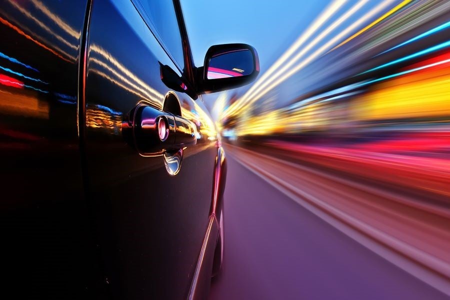 Side view of a dark car speeding at night with blurred streaks of colorful light behind it.
